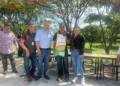 Grupo de agricultores y funcionarios en un evento al aire libre con árboles y mesas en el fondo.