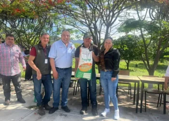 Grupo de agricultores y funcionarios en un evento al aire libre con árboles y mesas en el fondo.