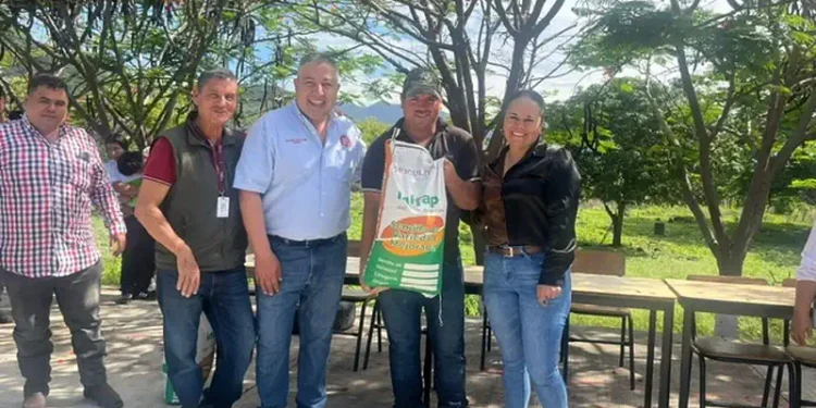 Grupo de agricultores y funcionarios en un evento al aire libre con árboles y mesas en el fondo.