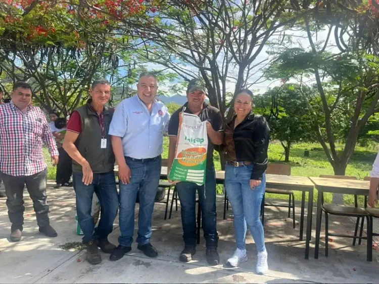 Grupo de agricultores y funcionarios en un evento al aire libre con árboles y mesas en el fondo.