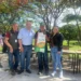 Grupo de agricultores y funcionarios en un evento al aire libre con árboles y mesas en el fondo.