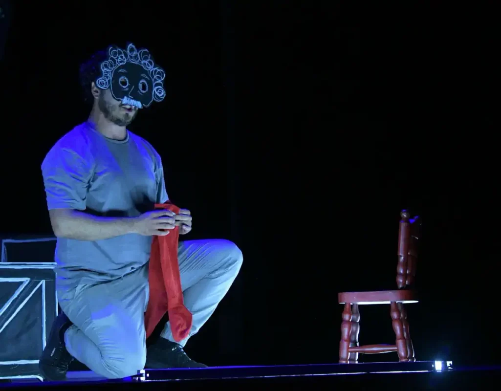 Actor con máscara de payaso y peluca en escena teatral oscura, sentado en el suelo con una silla y un telón de fondo negro.