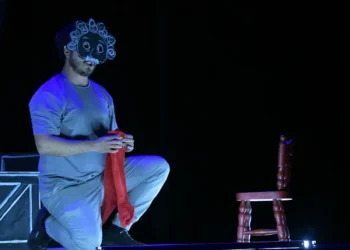 Actor con máscara de payaso y peluca en escena teatral oscura, sentado en el suelo con una silla y un telón de fondo negro.