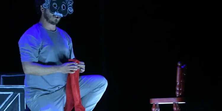 Actor con máscara de payaso y peluca en escena teatral oscura, sentado en el suelo con una silla y un telón de fondo negro.