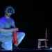 Actor con máscara de payaso y peluca en escena teatral oscura, sentado en el suelo con una silla y un telón de fondo negro.