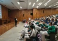 Grupo de estudiantes en uniforme de artes marciales asistiendo a una clase en un auditorio con madera en las paredes.