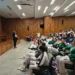 Grupo de estudiantes en uniforme de artes marciales asistiendo a una clase en un auditorio con madera en las paredes.