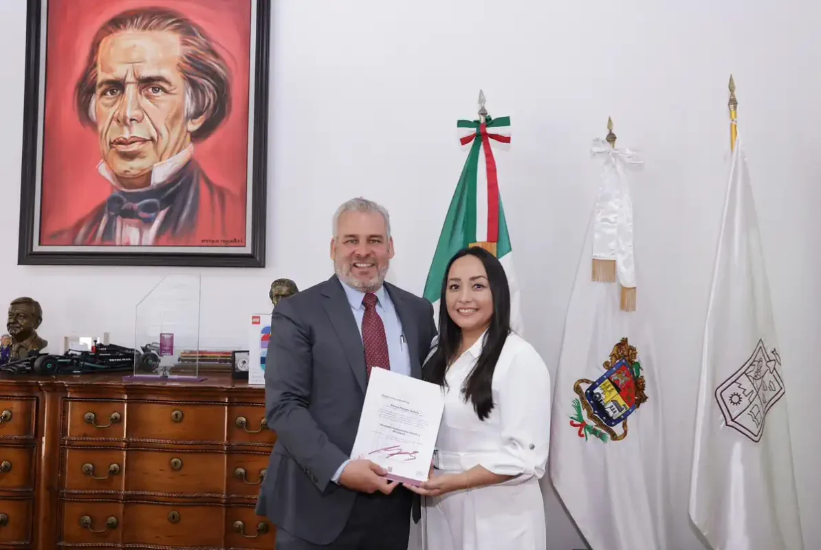 Hombre y mujer en oficina, entregando un documento, con retrato de un personaje histórico en la pared y banderas oficiales en.