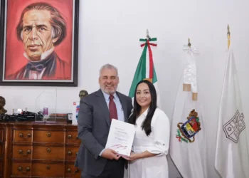 Hombre y mujer en oficina, entregando un documento, con retrato de un personaje histórico en la pared y banderas oficiales en.
