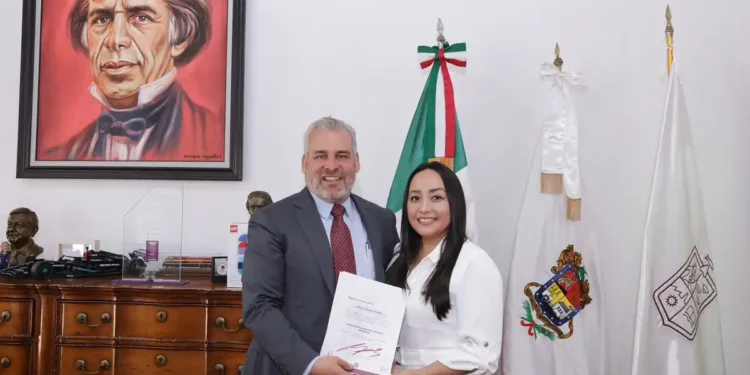Hombre y mujer en oficina, entregando un documento, con retrato de un personaje histórico en la pared y banderas oficiales en.