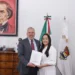Hombre y mujer en oficina, entregando un documento, con retrato de un personaje histórico en la pared y banderas oficiales en.