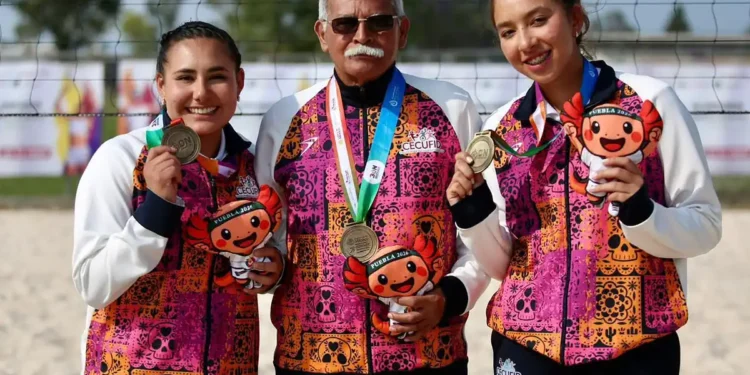 Dos jóvenes deportistas mexicanas y un entrenador posan con medallas en torneo de voleibol de playa en Jalisco.