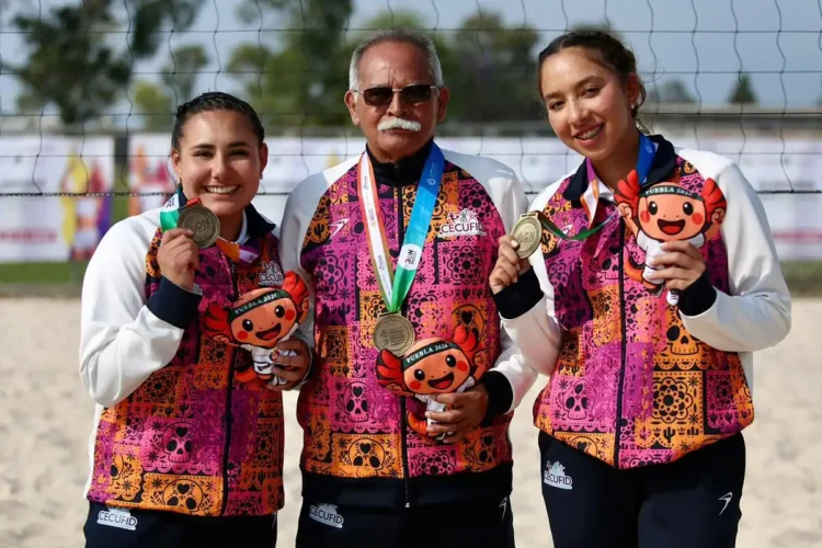 Dos jóvenes deportistas mexicanas y un entrenador posan con medallas en torneo de voleibol de playa en Jalisco.