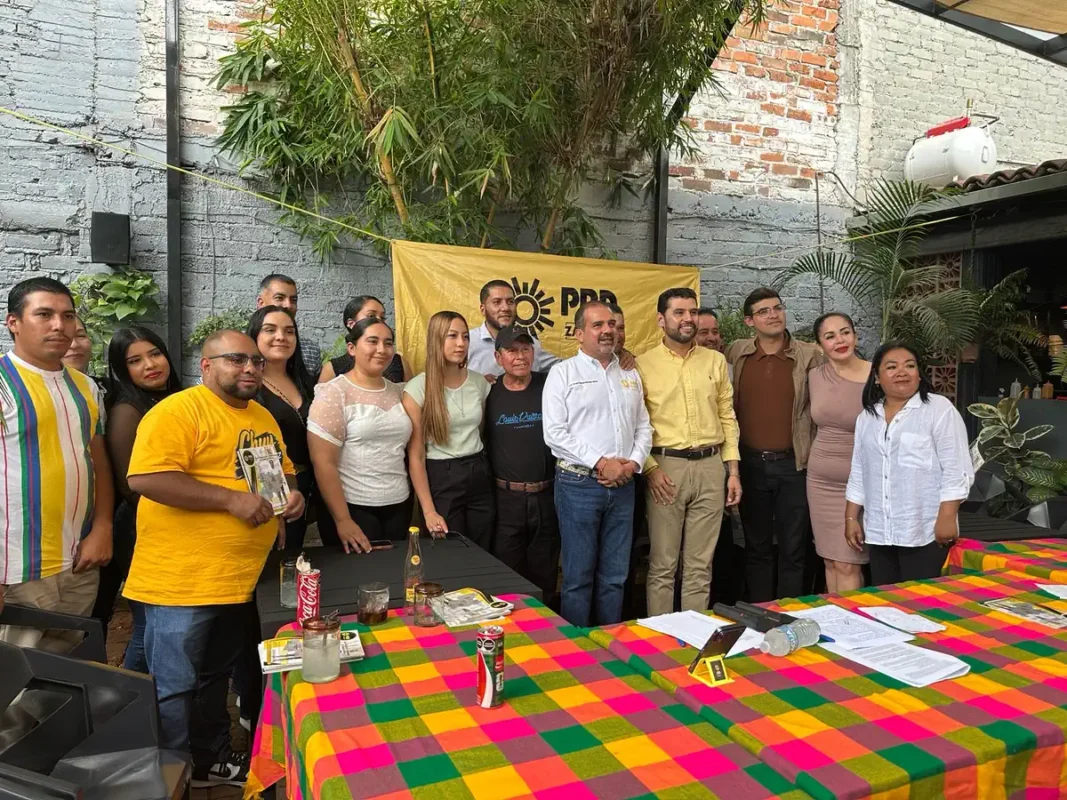 Grupo de personas en reunión política con bandera del PRD en fondo de muro de ladrillos y plantas.