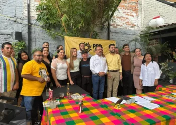 Grupo de personas en reunión política con bandera del PRD en fondo de muro de ladrillos y plantas.