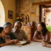 Familia conversando en una mesa de madera en una casa con paredes de piedra y ventanas grandes.