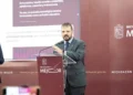 Hombre en traje oscuro hablando en conferencia en un escenario con fondo rojo y logotipo del gobierno de Michoacán.