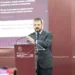 Hombre en traje oscuro hablando en conferencia en un escenario con fondo rojo y logotipo del gobierno de Michoacán.