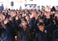 Niños y niñas en uniforme escolar levantando las manos en una actividad en un evento escolar.