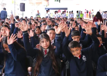 Niños y niñas en uniforme escolar levantando las manos en una actividad en un evento escolar.