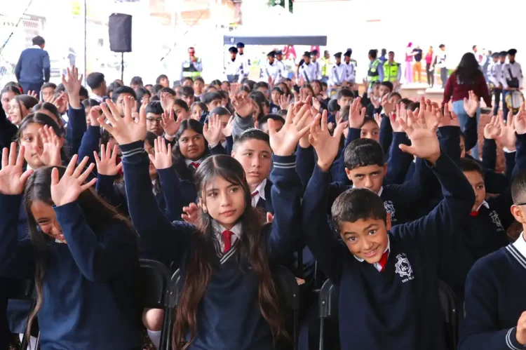 Niños y niñas en uniforme escolar levantando las manos en una actividad en un evento escolar.