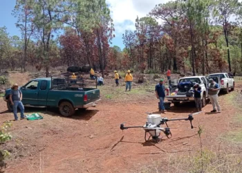 Drones sobre campo de tierra con vehículos y personas en Michoacán durante siembra de semillas.