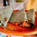 Tacos de tinga en tortilla de maíz en un plato de barro, con fondo difuso de puestos callejeros en Morelia.