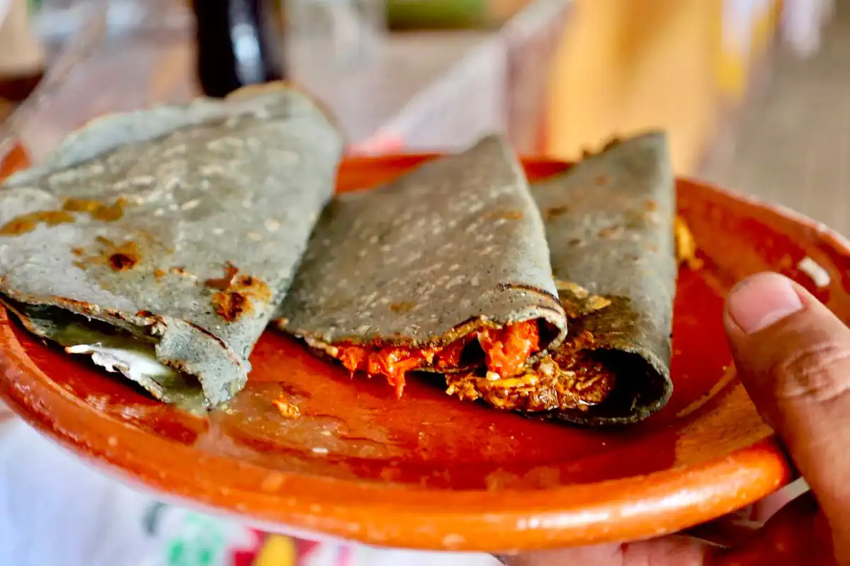 Tacos de tinga en tortilla de maíz en un plato de barro, con fondo difuso de puestos callejeros en Morelia.