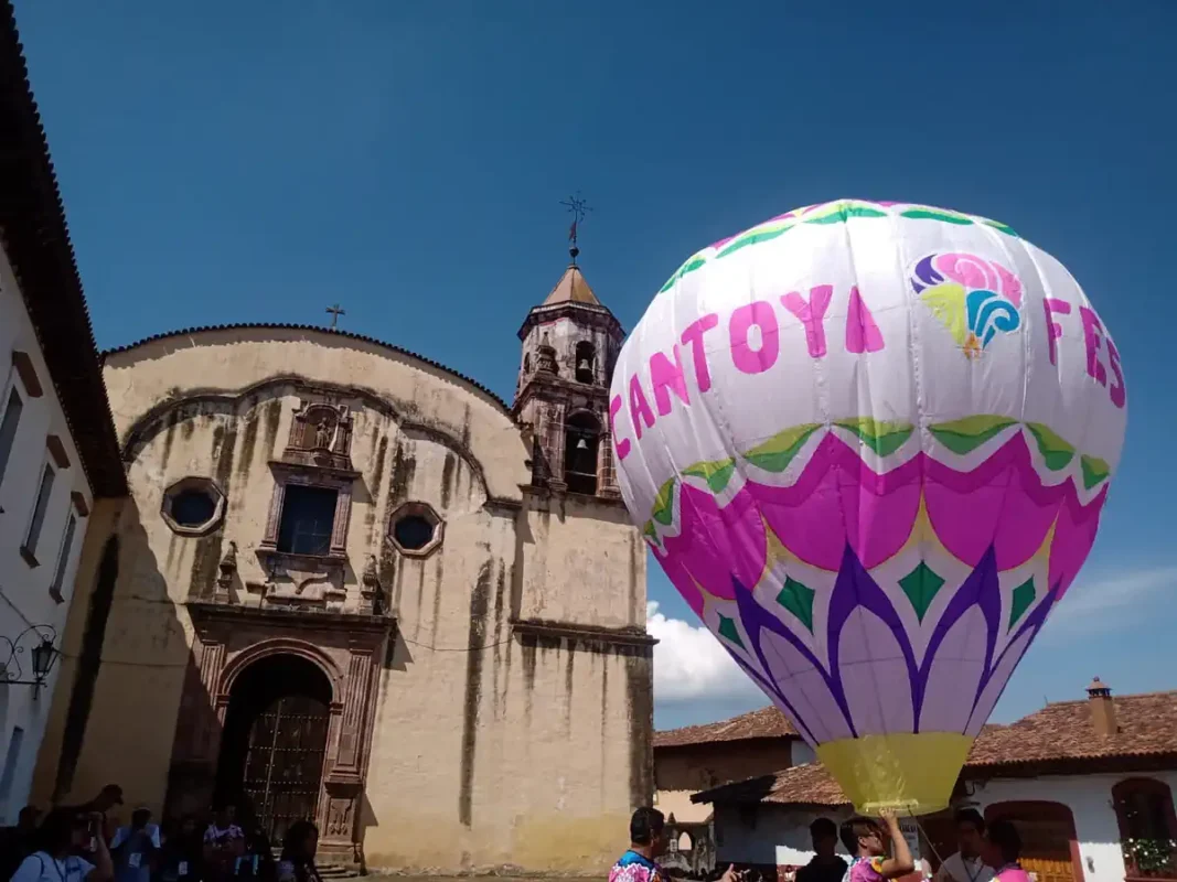 Globo aerostático con colores vibrantes y logotipo del Cantoya Fest en Pátzcuaro, con iglesia antigua de fondo y cielo despej.