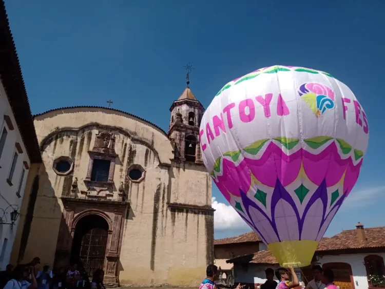 Globo aerostático con colores vibrantes y logotipo del Cantoya Fest en Pátzcuaro, con iglesia antigua de fondo y cielo despej.