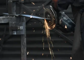 Trabajador usando amoladora para cortar metal en un entorno industrial, con chispas y luz tenue en el fondo.