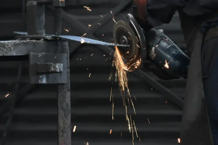 Trabajador usando amoladora para cortar metal en un entorno industrial, con chispas y luz tenue en el fondo.