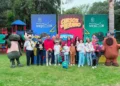 Niños y adultos disfrutan actividades en parque con personajes de caricaturas y banners promocionales de cursos de verano.