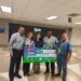 Grupo de cinco personas sosteniendo un cheque grande en un espacio interior, celebrando donativo récord de Aneberries.