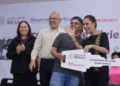 Cuatro personas en un evento oficial con fondo de logotipos del gobierno y patrocinadores, sonriendo y sosteniendo un cartel.
