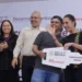 Cuatro personas en un evento oficial con fondo de logotipos del gobierno y patrocinadores, sonriendo y sosteniendo un cartel.