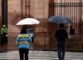 Personas con paraguas en la calle bajo lluvia en Michoacán, con fondo de edificio histórico y reja de hierro.