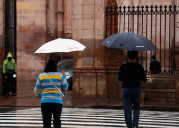 Personas con paraguas en la calle bajo lluvia en Michoacán, con fondo de edificio histórico y reja de hierro.