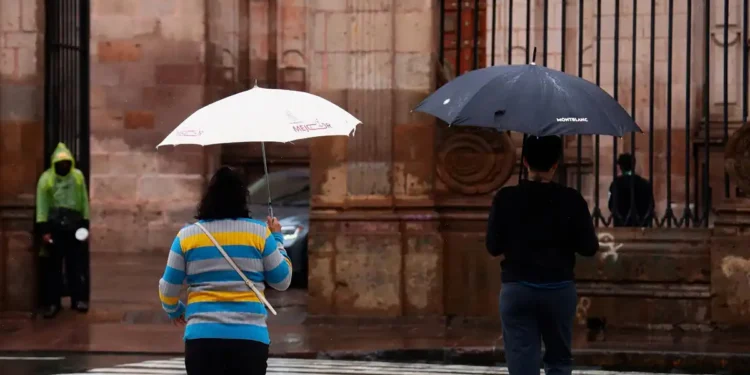 Personas con paraguas en la calle bajo lluvia en Michoacán, con fondo de edificio histórico y reja de hierro.