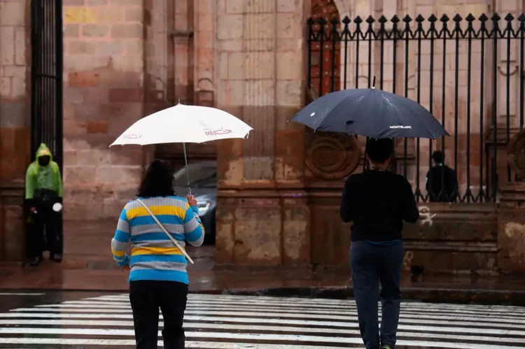 Personas con paraguas en la calle bajo lluvia en Michoacán, con fondo de edificio histórico y reja de hierro.