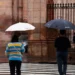 Personas con paraguas en la calle bajo lluvia en Michoacán, con fondo de edificio histórico y reja de hierro.