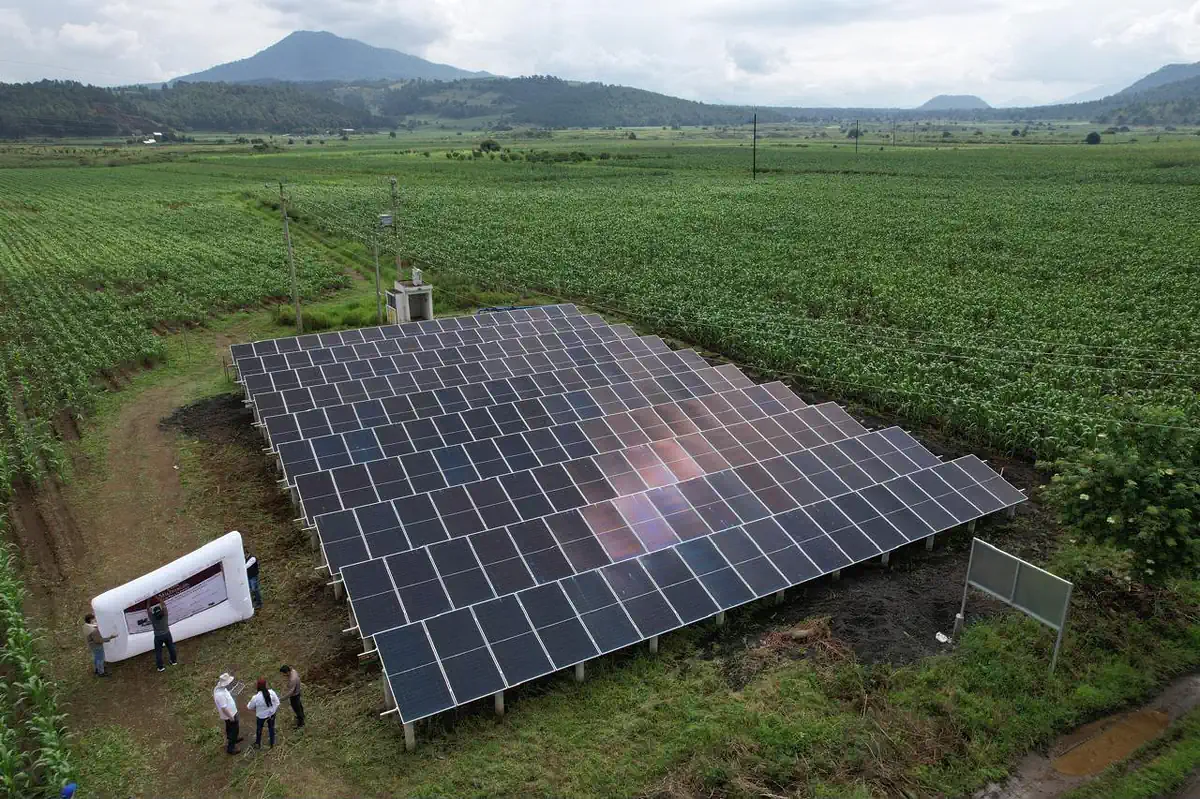 Paneles solares en campo abierto con montículo de tierra y fondo de montañas y cielo nublado.