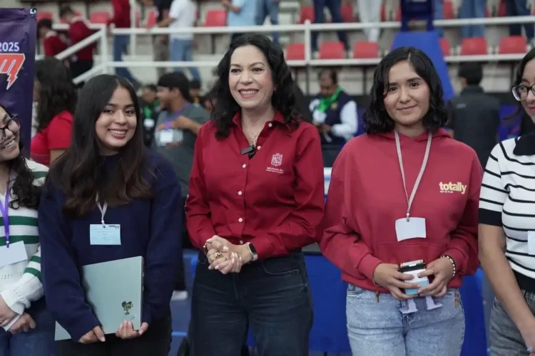 Mujeres jóvenes y una adulta en evento escolar, con fondo de auditorio y público en asientos rojos y blancos.