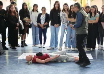 Estudiantes observan un simulacro de primeros auxilios con un maniquí en un gimnasio escolar.