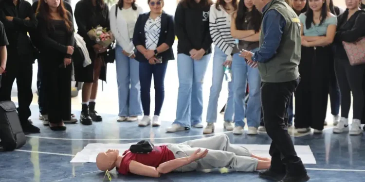 Estudiantes observan un simulacro de primeros auxilios con un maniquí en un gimnasio escolar.