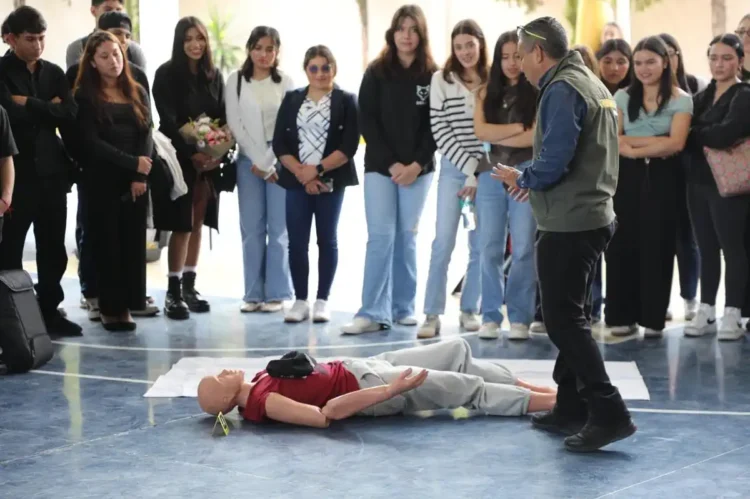 Estudiantes observan un simulacro de primeros auxilios con un maniquí en un gimnasio escolar.