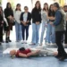 Estudiantes observan un simulacro de primeros auxilios con un maniquí en un gimnasio escolar.