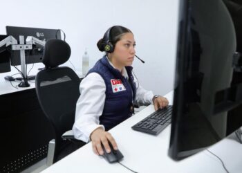 Mujer en centro de atención telefónica con auriculares y computadora, monitoreando llamadas para prevenir extorsiones.