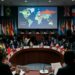 Delegados de diferentes países en una conferencia global con banderas y mapa mundial en pantalla.