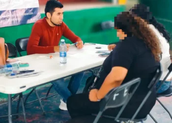 Joven realiza toma de muestras de ADN a mujer en evento en Sahuayo, con fondo de escaleras y banner informativo.
